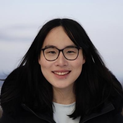 Smiling young woman with eyeglasses and long dark hair, wearing a black jacket.