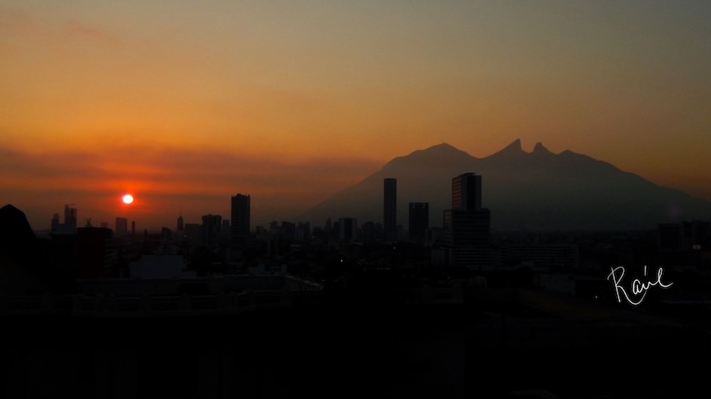 Distant sunset, small pink sun in dark yellow horizontal clouds, with a city and mountains in the foreground.