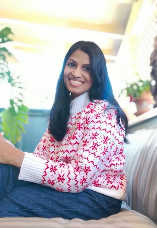 Smiling young woman with long dark hair wearing a white sweater with red snowflake pattern.