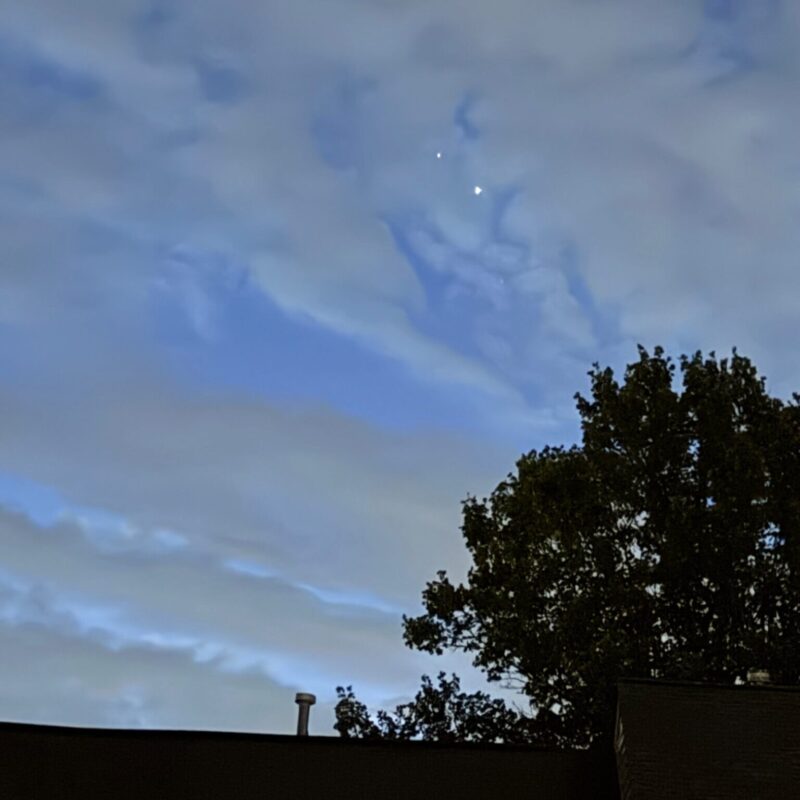 Two bright dots in a cloudy sky, above the roof of a house and a tree.