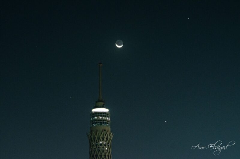 Lit top of an ornate tower with the moon and 2 bright dots to its top right.
