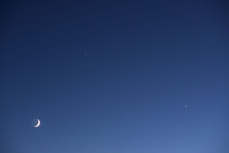 Blue sky with a thin moon at the bottom left, a dot to the top and another one to the right side.