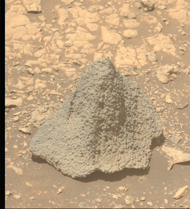 Conical rock on brownish, sandy terrain, looking like a cover is draped over it.