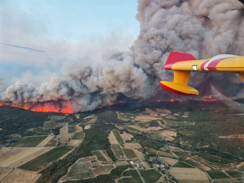 Wildfire in France: Part of a plane and looking down on the landscape with green and brown and a bit of bright orange with billowing smoke clouds above.