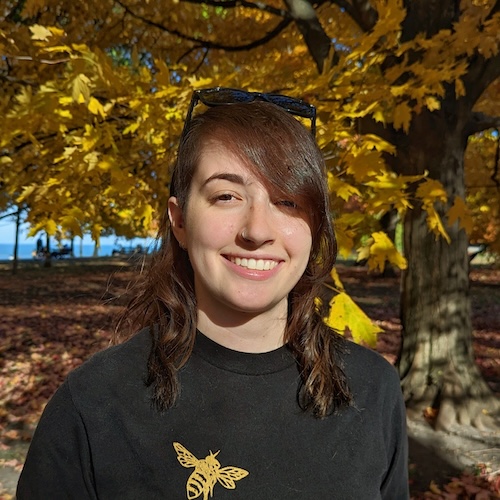 Smiling young woman with long brown hair standing under a tree. The distant ocean is behind her.