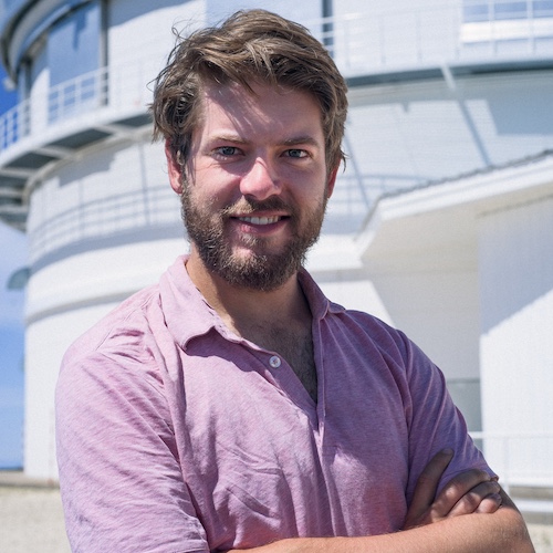 Smiling man standing with his arms folded, with a white curved building behind him.