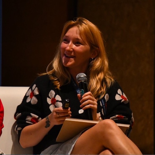 Smiling woman with long blond hair sitting in a chair and holding a microphone. A laptop is also in her lap.