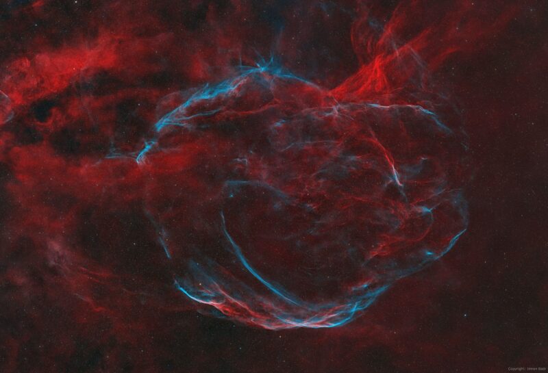 Large, arc-shaped clouds of gas in red and blue, with dark areas and numerous background stars.