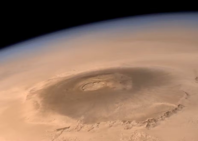 Oblique orbital view of a huge sprawling volcano with a big crater on reddish planet.