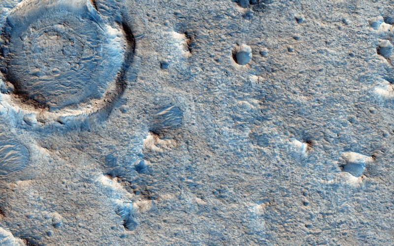 Overhead view of relatively flat terrain with a few small knobs of rock and a crater in the top left corner.