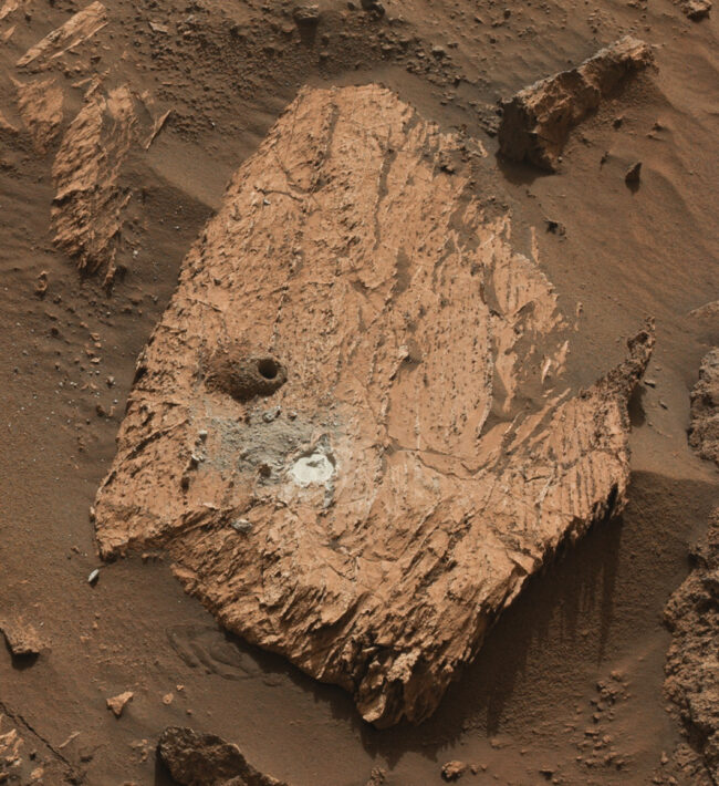 Mars rock: An arrowhead-shaped rock, orangish with some black markings, a drilled hole and a white spot.