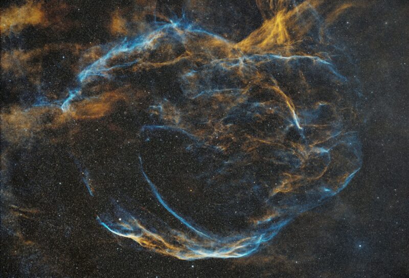 Large, arc-shaped clouds of gas in blue and orange, with dark areas and numerous background stars.