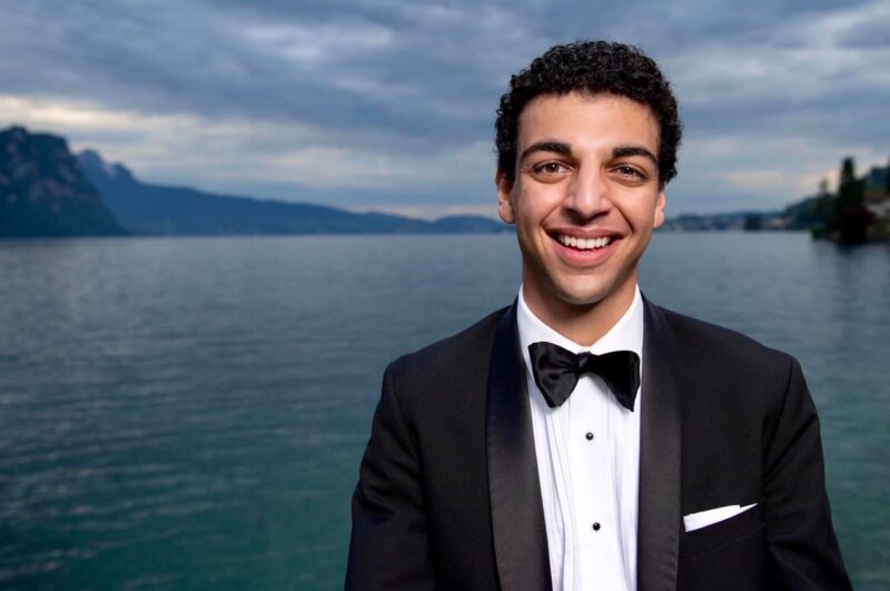 Planet Y: Young man with short dark hair, in formal dress, standing in front of a body of water.