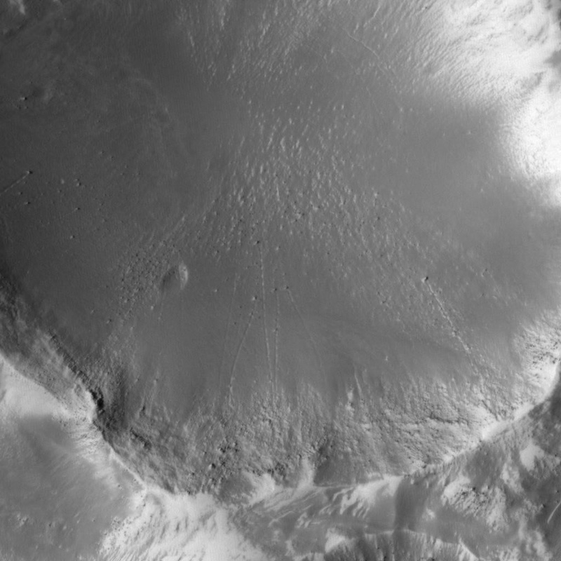 Black and white view from above of part of a crater. Small stones have rolled down from the outer edge, leaving trails behind them in the sand.