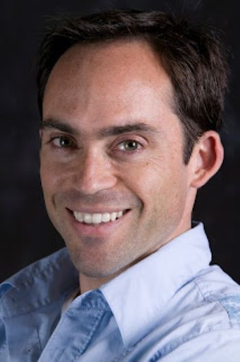 A man with dark hair and a blue button-up shirt smiles at the camera.