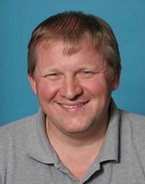 Smiling man with short hair wearing a gray shirt, on blue background.