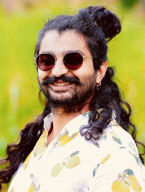 Smiling man with beard, long dark hair, hair bun and sunglasses, wearing a bright shirt with lemons printed on it.