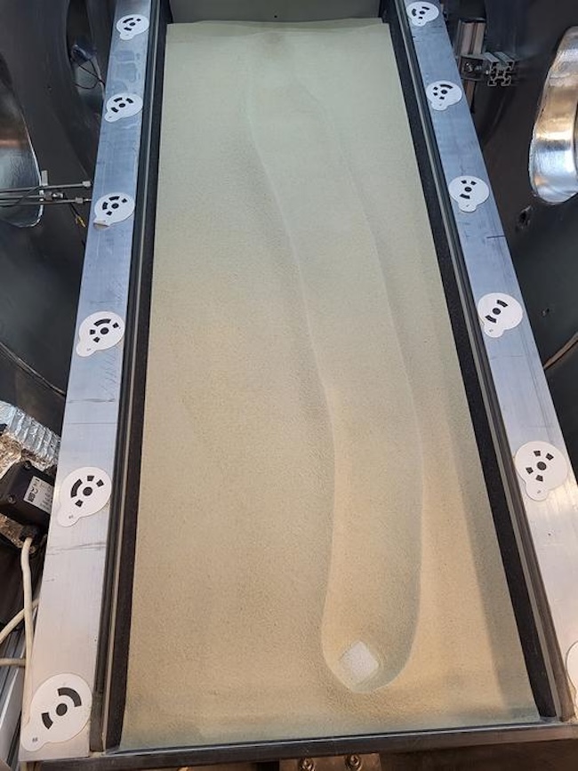 Long rectangular metal container with sand in it. A white chunk of ice has moved lengthwise, leaving a trail behind it.
