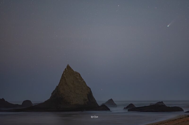 A beach with large rocks sticking up and a comet in the sky.