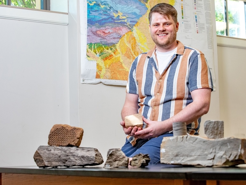 Smiling young man wearing a striped shirt with a table covered in rocks, and holding a rock.