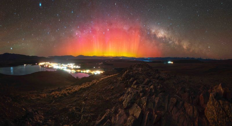 Looking down from a mountain on a lit up town with a splendid orange red aurora beneath the arc of the Milky Way.