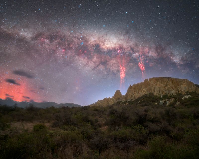Rugged mountains with cloudy band of Milky Way above, 2 red sprites in front of the Milky Way, and a red glow to the left.