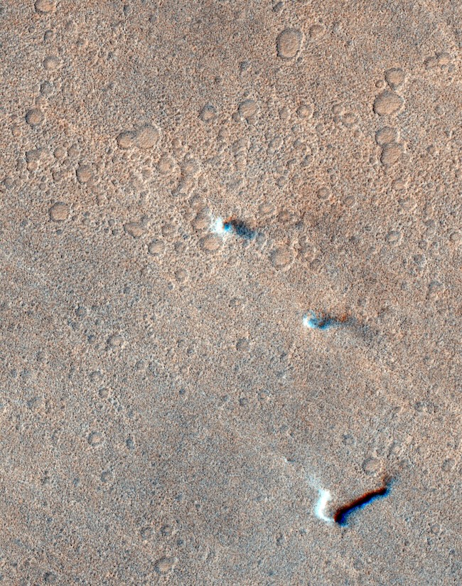 View from above of 3 small vertical clouds on a cratered surface.