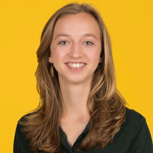 Smiling woman with long hair wearing a dark shirt, on yellow background.