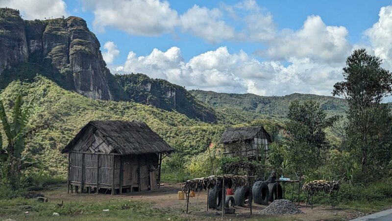 Wood huts, drying racks, a few trees beneath lowering cliffs. Above, a blue sky with some white clouds.