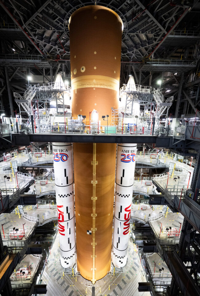 A large orange cylinder and two rocket boosters inside a huge building.