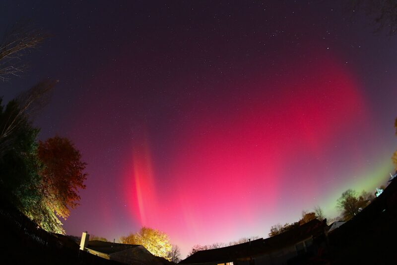 A wide-angle view at night with red and green light in the sky and a house in the foreground.