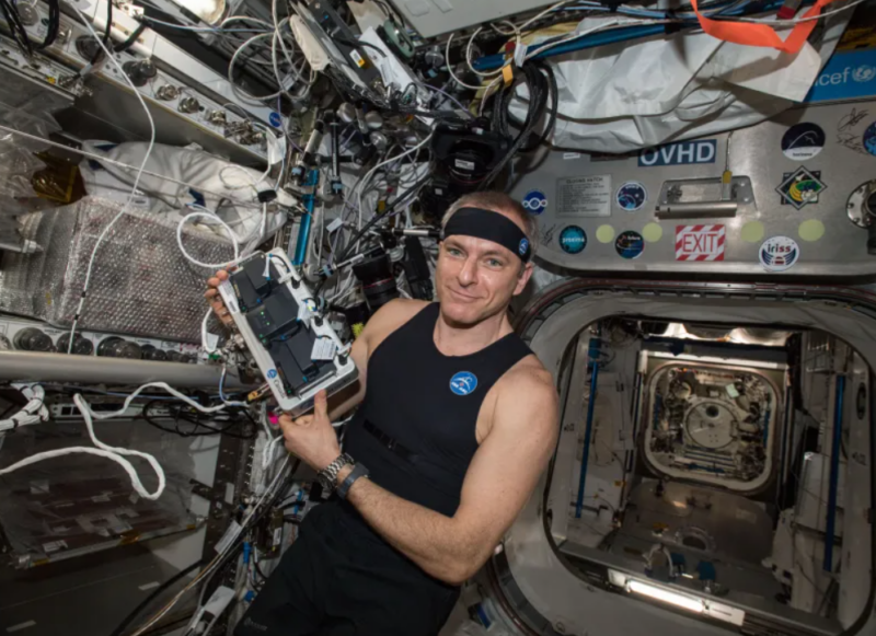 A man in a tank top holding a device while on the space station.
