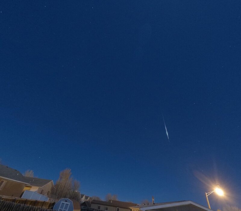 A thin white streak slightly bulging in the middle in a dark blue sky, above rooftops.