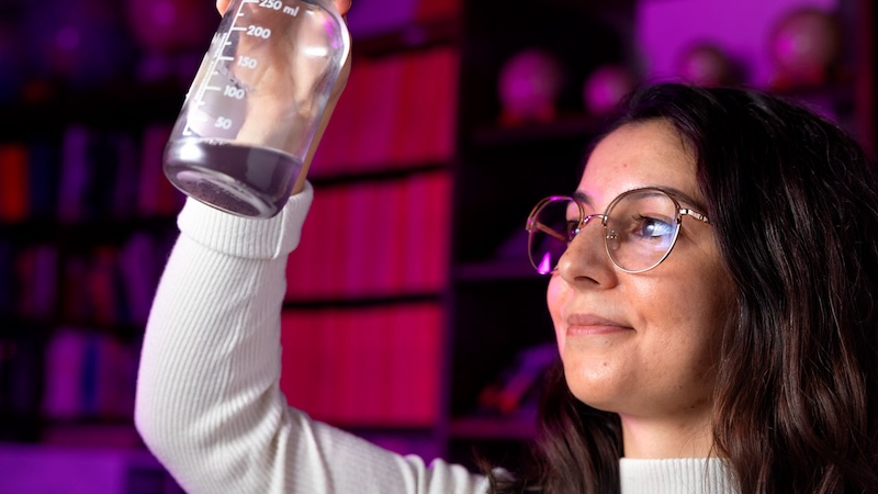 Smiling young woman with eyeglasses and dark hair holding up a flask with some dark liquid in it.
