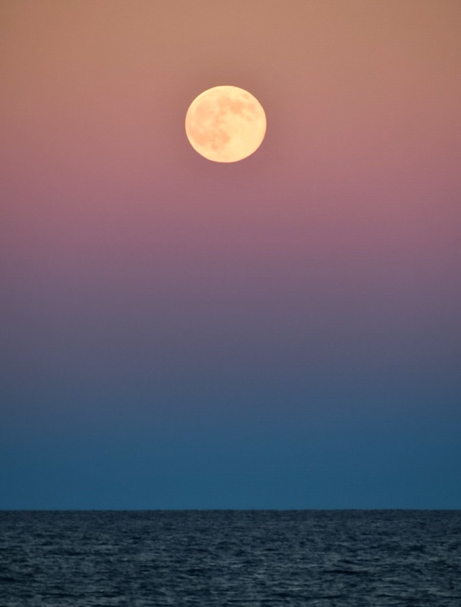 Bright full moon in pink sky above dark blue fuzzy band at the ocean horizon.