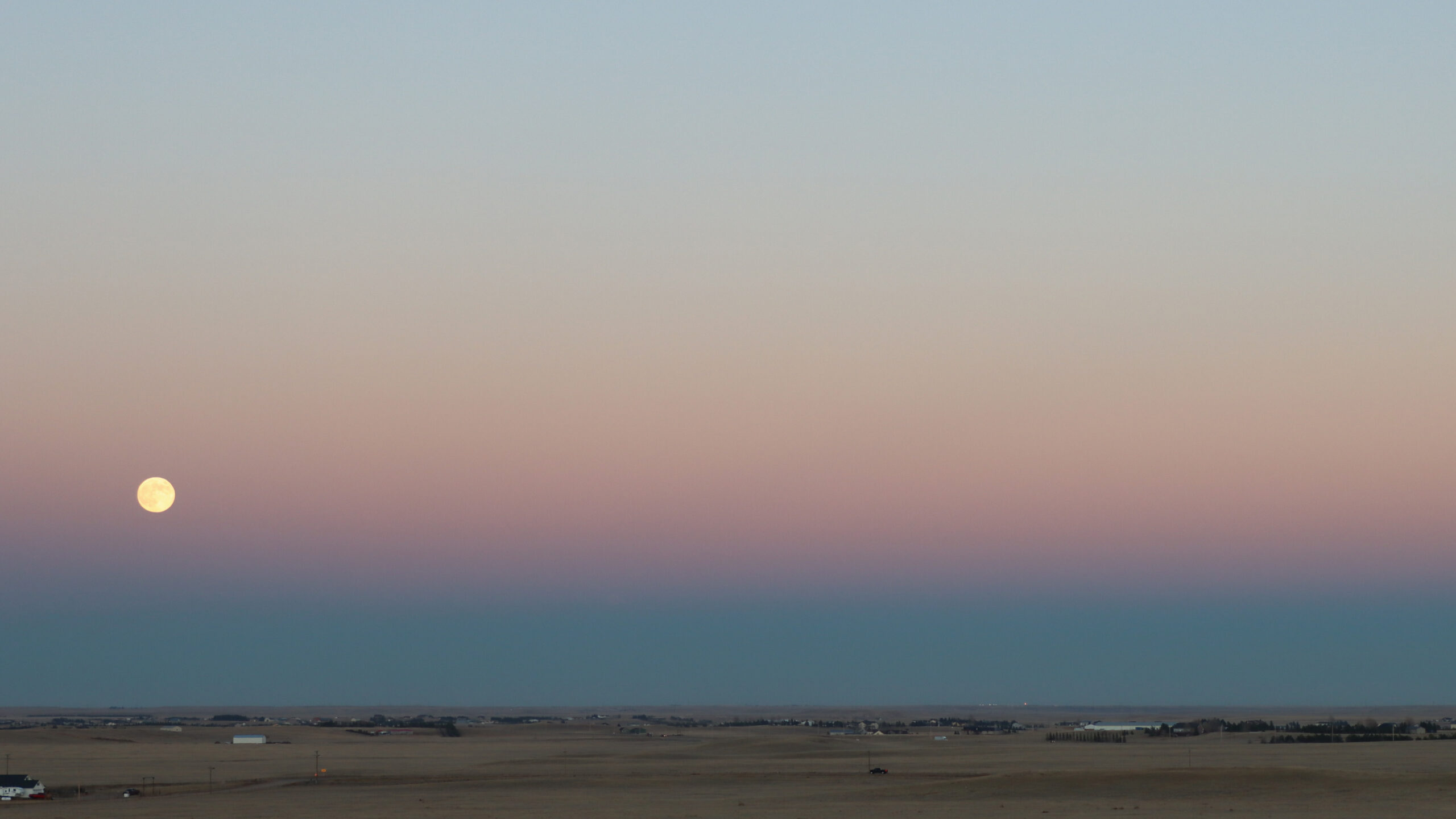 A full supermoon lying within a pink band above a dark blue band on a distant horizon.