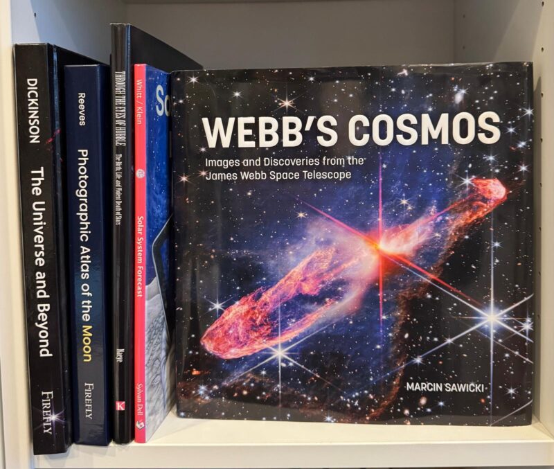 Webb's Cosmos: Front cover of a book on a bookshelf with other astronomical picture books, with a space photo and the title Webb's Cosmos.