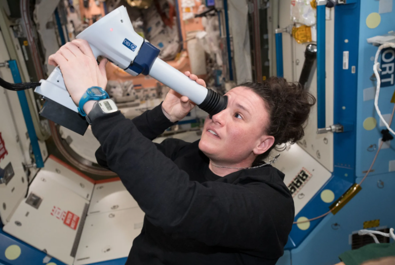 A woman floating in the space station with a large projectile type machine held up to her eye.