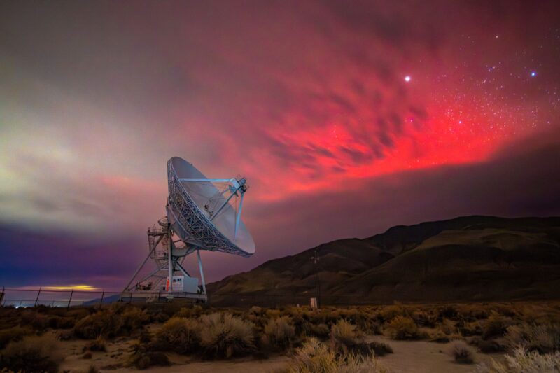Radio telescope with the shape of a cone located in an open field. The sky looks deep red and there are many bright stars.