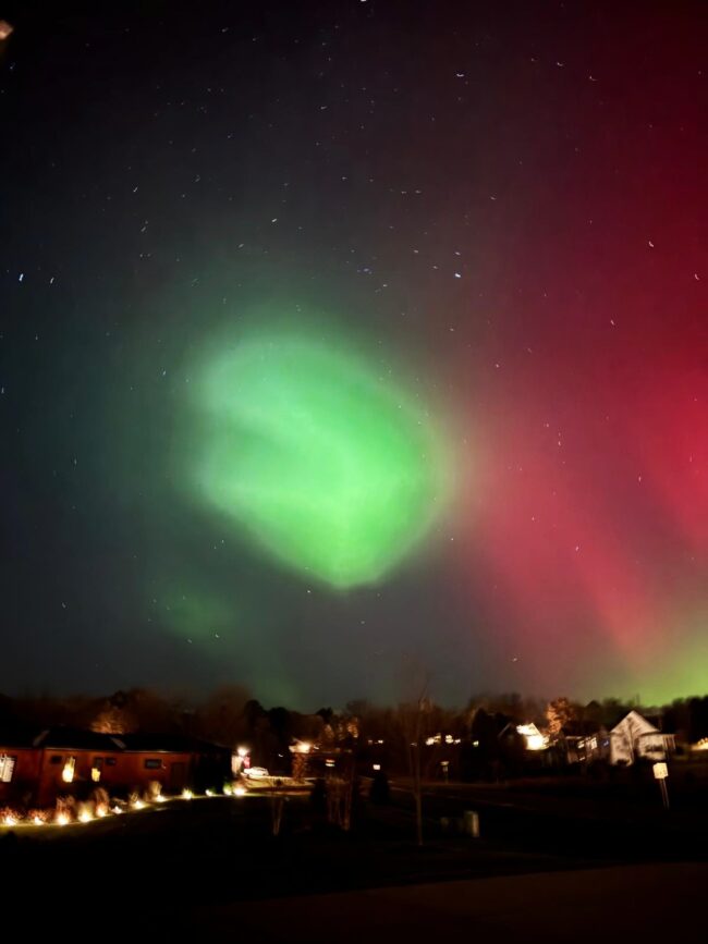 Reddish green aurora at right with a bright blob of green at center and some lit houses below.