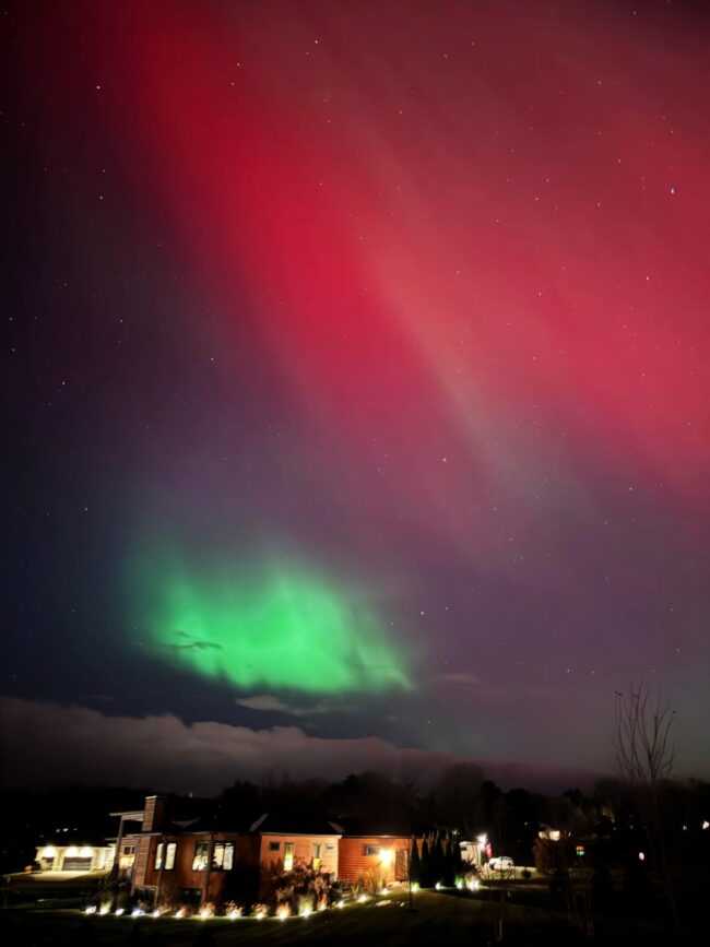 Reddish aurora with a bright green blob lower to the horizon and above clouds and a house with a lit perimeter.