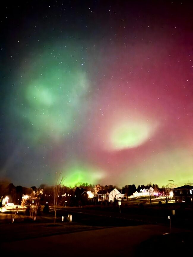 Reddish green aurora with bright green blobs in the foreground and a few lit houses below.
