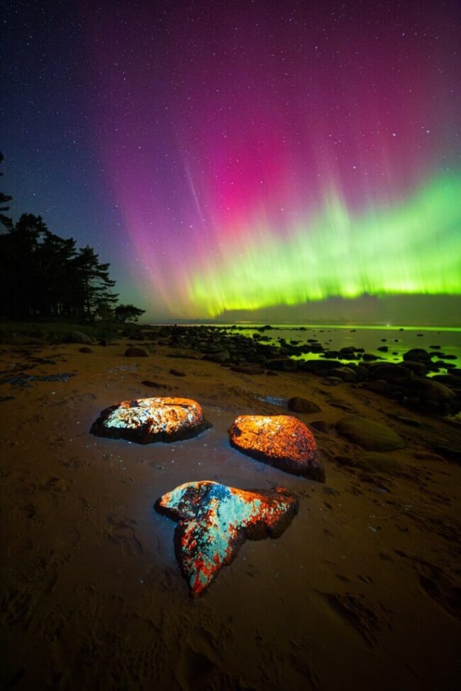 Shiny rocks on the shore with greenish yellow and red aurora behind.