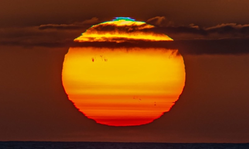 Close-up of the sun at sunset, a bit distorted with clouds in front of the sun and a blue and green flash on top.