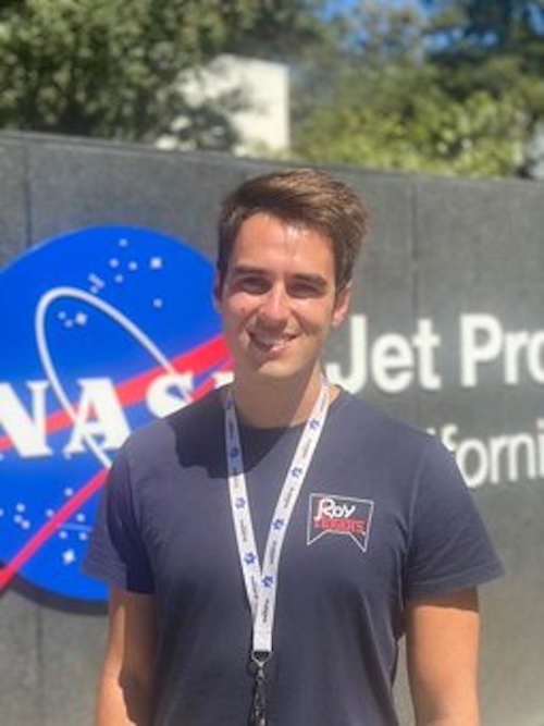 Smiling man with a lanyard around his neck. The NASA logo is on a cement wall behind him.