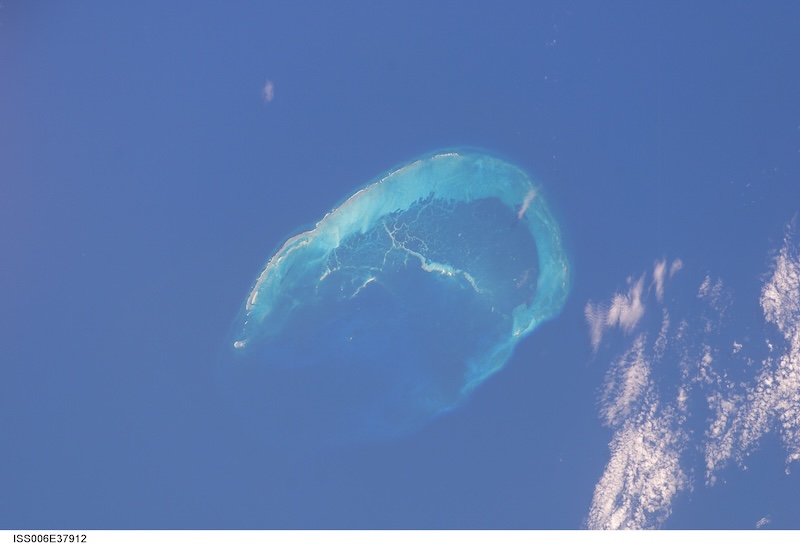 Плави океан са атолом у облику полумесеца и мало облака.