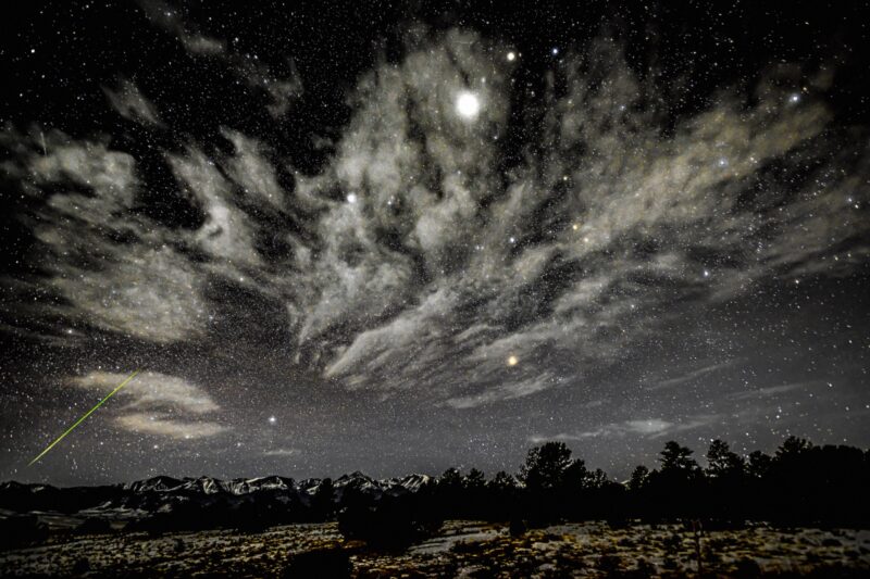 Dark sky covered in countless stars. There are some clouds and a bright, green streak at the bottom left.