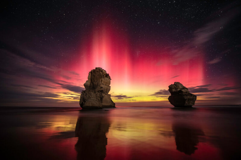 Red and yellow aurora behind large pillars of rock on the beach.