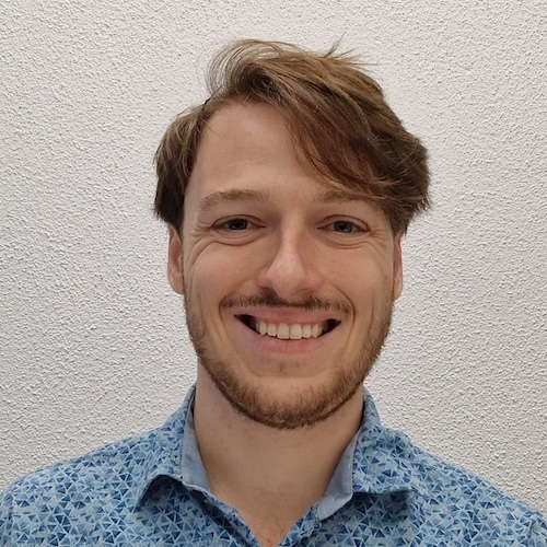 Насмејани човек са таласастом косом и подшишаном брадом и брковима, носи плаву кошуљу са узорком.