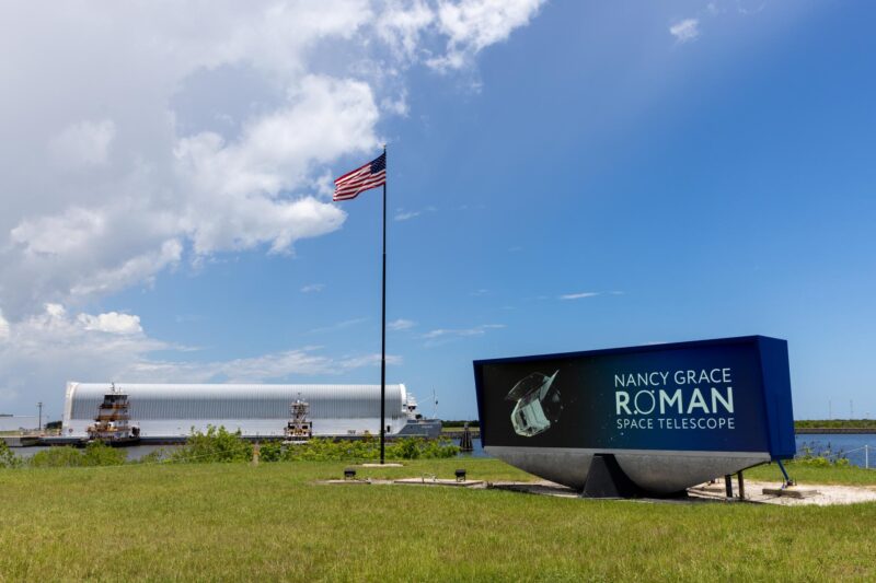 A large white tube at harbor with American flag and a sign for the Nancy Grace Roman telescope.